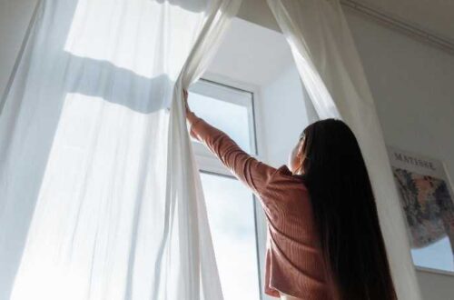 Woman opens white curtains in bright room, revealing a sunny day outside., tags: kosten im - pexel