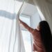 Woman opens white curtains in bright room, revealing a sunny day outside., tags: kosten im - pexel