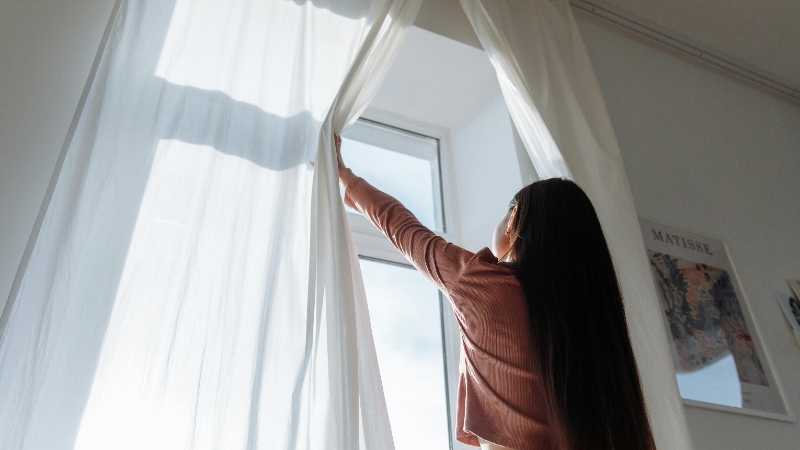 Woman opens white curtains in bright room, revealing a sunny day outside., tags: kosten im - pexel